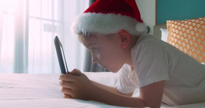 Curious happy cute smiling child in Christmas hat Santa Clausa, kid boy using digital tablet technology device lying on bedroom alone. Small child hold pad computer surfing internet play game at home