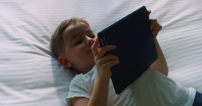 Cute smiling child, kid boy using digital tablet technology device lying on bedroom alone. Small child hold pad computer surfing internet play game at home