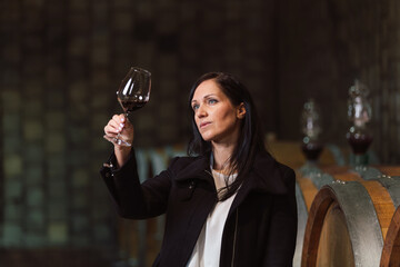 Woman in the wine cellar with barrels in background drinking and tasting wine.