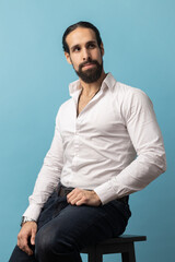 Obraz premium Unsmiling serious dark haired attractive man with beard wearing white shirt and black trousers, looking away with attentive look while sitting on chair. Indoor studio shot isolated on blue background.