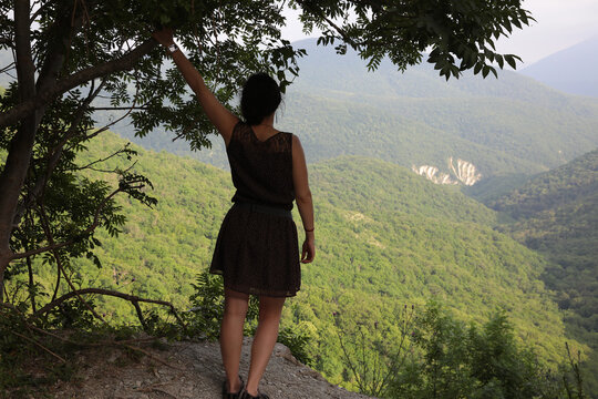 A girl standing on the edge of a cliff holds her hand to a tree and admires the landscape of the tops of mountains covered with forest.Back view full length image of female traveler