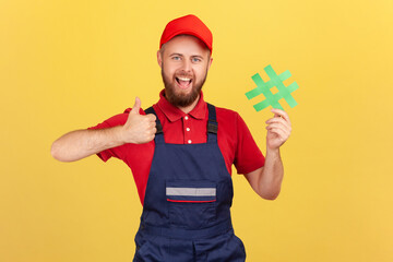 Portrait of excited handyman wearing blue overalls standing holding green hashtag and showing thumb up, looking at camera with smile. Indoor studio shot isolated on yellow background.