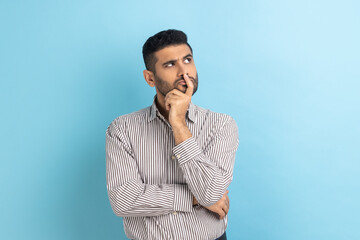 Portrait of thoughtful handsome bearded businessman holding his chin and pondering idea, confused not sure about solution, wearing striped shirt. Indoor studio shot isolated on blue background.