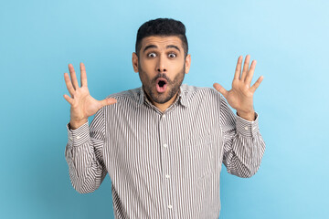 Fototapeta premium Portrait of surprised astonished bearded businessman standing with open mouth and raised arms, being impressed of shocking news, wearing striped shirt. Indoor studio shot isolated on blue background.