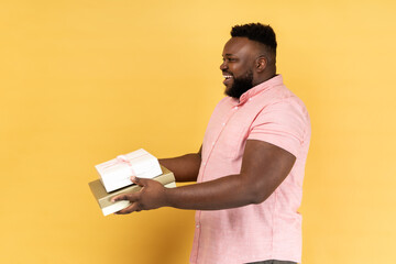 Side view portrait of satisfied happy man wearing pink shirt holding giving present boxes, congratulating, looking ahead with toothy smile. Indoor studio shot isolated on yellow background.