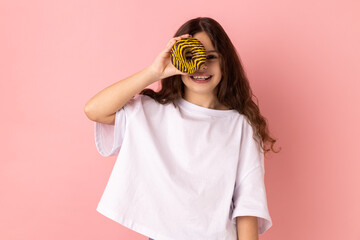 Portrait of funny adorable little girl wearing white T-shirt standing having fun, holding and covering eye with donut and smiling. Indoor studio shot isolated on pink background.