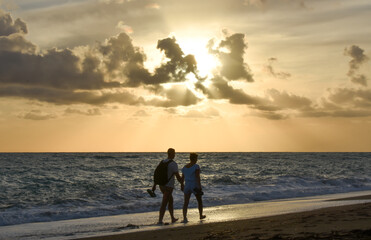 Young couple hand to hand walking on a beach at cloudy sunset