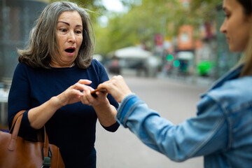 Street theft concept. Young female thief trying to snatch mobile phone of frightened elderly woman...