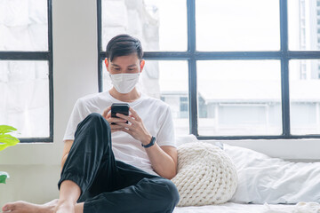 Asian casual man wear medical mask safety health using smartphone online sitting on white bed at home. New normal male with face mask use mobile phone in morning. Lifestyle technology self isolation.
