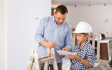 Friendly adult man with construction documents talking with preteen son and gesturing inside apartment being renovated