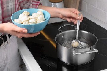 Woman putting frozen dumplings into saucepan with boiling water on cooktop in kitchen, closeup