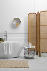Obraz na płótnie Canvas Modern ceramic bathtub and table with toiletries near white wall in room