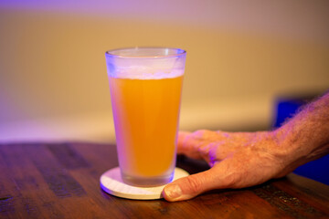 A tall glass of cold craft beer with a thick head of white froth. The Irish red ale beer glass is on the edge of a wooden table at a brewery. The gold alcohol refreshment beverage of draft beer.    