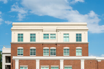 The top three floors of a brown brick apartment building with multiple green tinted glass windows and a cream colored cement top floor. Behind the building, there's a blue sky and clouds.
