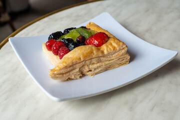 A square fruit filled French puff pastry on a white square plate. The cream and custard filled dessert has strawberries, kiwi, and blackberries on top with a sugar glaze. The pastry is flaky.	