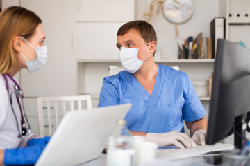 Obraz premium Adult doctor in medical face mask working on laptop in office, consulting with female colleague about patient diagnosis