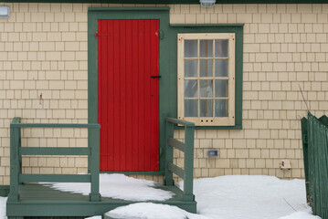 A single red wooden door with a vintage latch. There's a single window with multiple glass panes and green trim. The exterior surface of the wall is made of cedar shakes and painted pale yellow color.