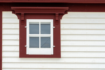 The exterior of a vintage white wooden house with horizontal textured clapboard and dark red trim. There's a small old four pane glass window with thick dark red trim. The eave is dark red. 