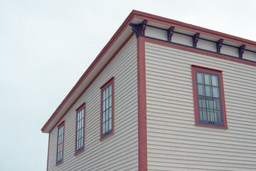 The roof section of a large white vintage wooden house with decorative pink and purple wood trim. There are three multi-pane windows on the top floor. The background is a dramatic blue cloudy sky.