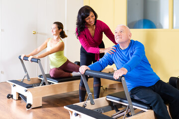Focused positive elderly man practicing pilates system on reformer to improve and maintain mobility under supervision of qualified Hispanic female trainer