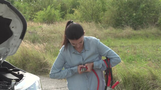 Emergency stop of car. Beautiful young weak woman unsuccessfully tried to open the wire alligator clips of starting cables. She's upset, stand alone on the side of deserted road, look around for help.