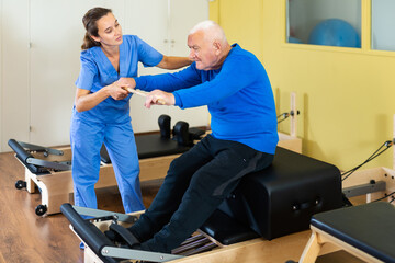 Qualified female physiatrist helping focused elderly man exercising on pilates reformer with wooden stick. Therapeutic gymnastic and rehabilitation concept