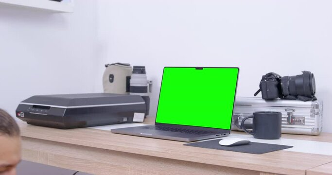 Adorable dachshund dog in shirt with jacket and with cap on its head stands on chair in front of laptop and barks, then leaves, view from the back. Green chromakey on gadget screen.