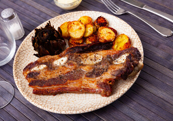 Delicious grilled beef ribs served on plate with side dish of fried potatoes and baked artichokes. Popular Spanish combination meal with churrasco