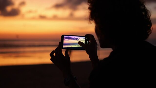 Silhouette of a woman's head with shoulders against the backdrop of a twilight sunset. A woman holds a smartphone in her hands and takes pictures of the sun hiding behind the horizon.