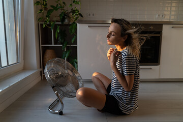 Exhausted young woman refreshing sit near big indoor ventilator blowing cooling fresh air at home on floor bonding. Tired female enjoying conditioner having fun indoors. Summer, heat weather concept - obrazy, fototapety, plakaty