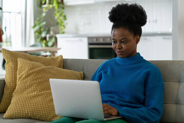 Fototapeta na wymiar Young African American woman freelancer typing article for online magazine in laptop sits on sofa in modern apartment. Black girl with fluffy hair performs remote work in comfortable environment