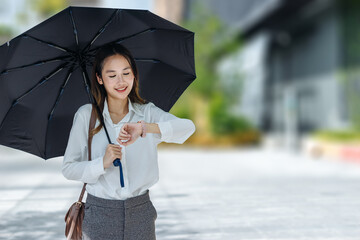 Cute Asian business woman with holding black umbrella looking at watch outdoors on sunny day, focus to the watch