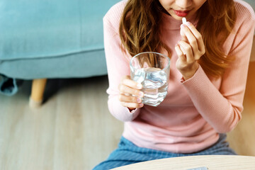Sick Asian woman eating pills with a glass of water in hand near window in her house. Close up to a glass of water.