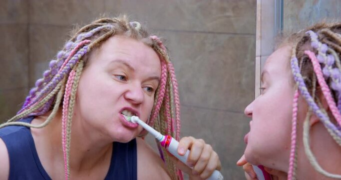 Swollen woman with hangover brushing her teeth thoroughly with electric toothbrush in morning after party. Lesbian with bright colored de braids stands in bathroom in front of sinks morning routine