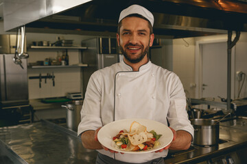 Handsome chef of restaurant showing plate with cesar salat standing on kitchen. Ready to eat