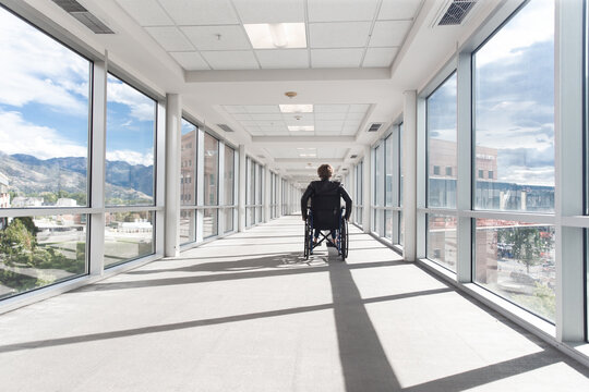 Solitary middle aged woman living with disability in a wheelchair wheeling herself down a hallway in a medical center or other building on a sunny day