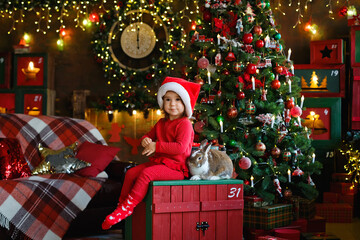 A cute little girl in a Santa hat and red pajamas is sitting next to a rabbit at a Christmas tree with bokeh lights. Pet care during the Christmas holidays.