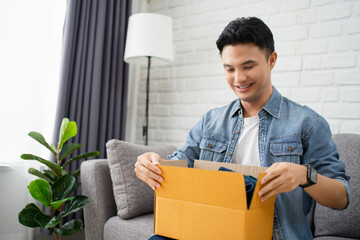 Young Asian man unpacking received parcel. He is sit on couch at home satisfied with Internet order shopping online, delivery service concept.