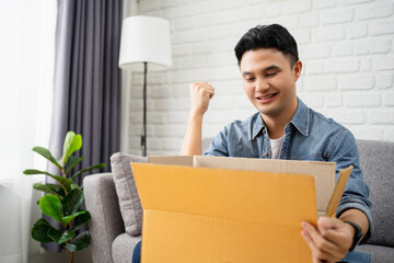Young Asian man unpacking the parcel paper box. He's making a winning gesture and satisfied with...