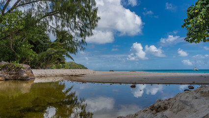 A calm pond on a tropical island. Lush green vegetation on the shore. The turquoise ocean is visible in the distance. Clouds in the blue sky. Reflection. Seychelles. Praslin. Anse Lazio beach  