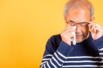Asian elder man crying raise glasses with tissue wipe red eyes studio shot isolated on yellow...