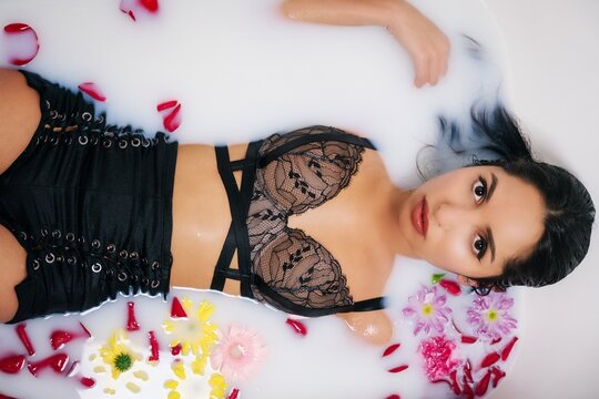Woman wearing black clubwear sitting in a tub of milk and flower petals