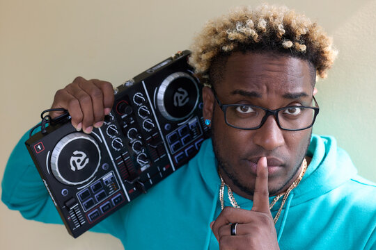 Dark skinned guy with a serious expression in a blue hoodie holds a DJ remote on his shoulder. Young black man in glasses with an Afro hairstyle presses his finger to his mouth, calling for silence.
