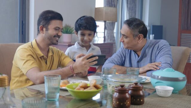 A happy Indian Ethnic young kid boy or grandson, son, and grandfather watching a funny video together on a mobile phone or a smartphone at the dining table before lunch or a meal in an indoor home.
