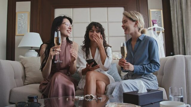 Young bride and her bridesmaids sitting on couch in living room holding glasses with sparking wine taking selfie photos on smartphone