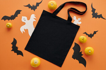 Top-down view shot of flat lay composition with black shopper bag, small pumpkins and paper bats and ghosts on orange background