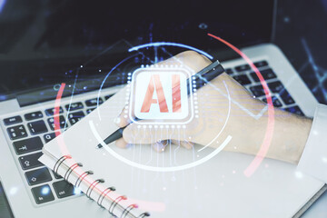 Double exposure of creative artificial Intelligence abbreviation with hand writing in notepad on background with laptop. Future technology and AI concept