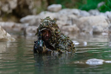 A military man or airsoft player in a camouflage suit sneaking the river and aims from a sniper...