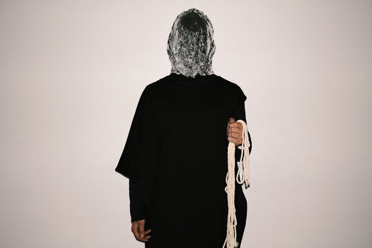 Horizontal medium studio portrait of unrecognizable strange man wearing black clothes with aluminum foil mask standing against white background holding rope