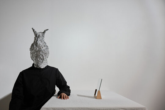 Horizontal studio shot of unrecognizable African American fashion model wearing black outfit with weird aluminum foil mask on head sitting at table
